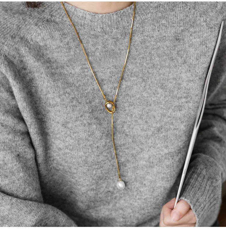Person wearing a gold necklace with a pearl pendant on a gray sweater.
