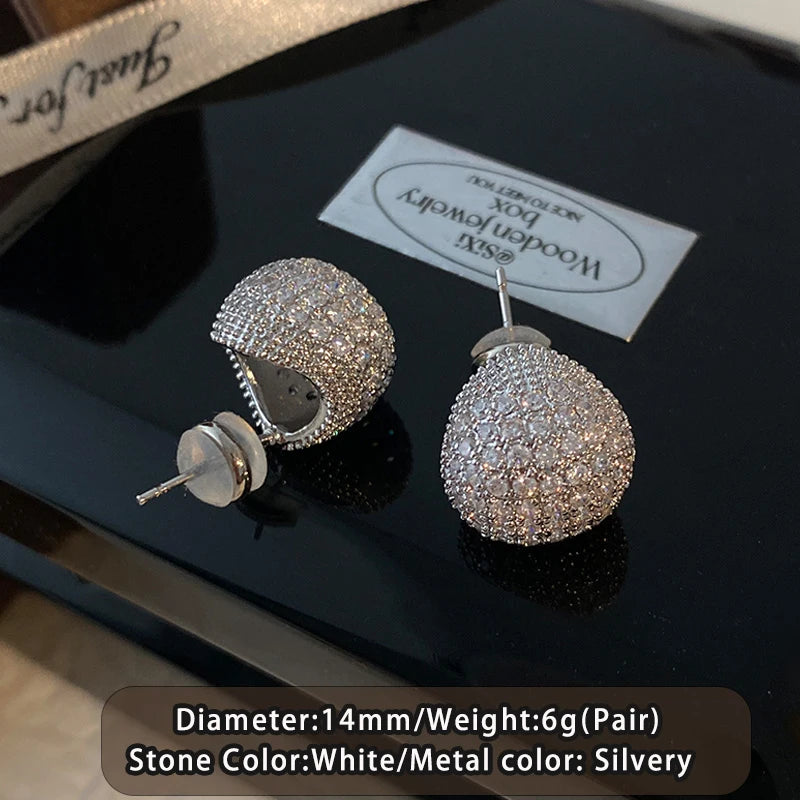 Pair of silver earrings on a black surface with a brand label in the background.