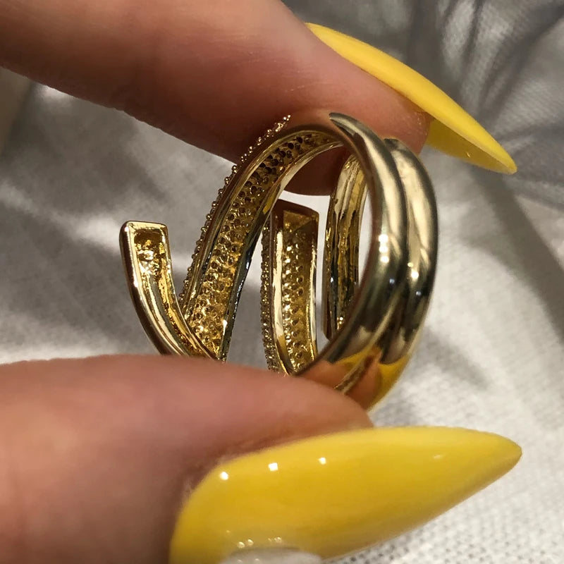 Gold hoop earrings held between fingers with yellow nail polish on a light background