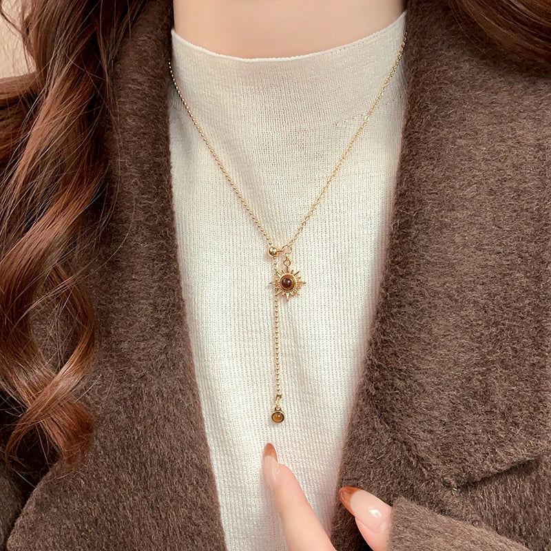 Person wearing a gold necklace with a sun pendant, pointing to it.