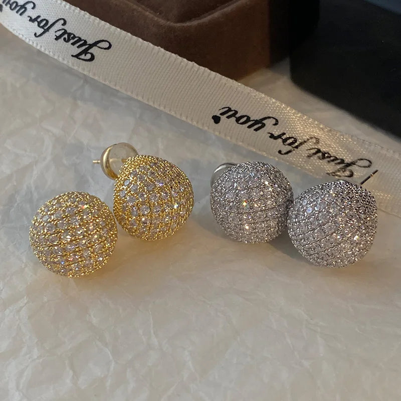 Pair of gold and silver ball-shaped earrings on a textured surface with a ribbon in the background.